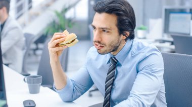 Genç İşadamı Masaüstü Bilgisayarında Oturan Hamburger Yiyen, Çöreği Çiğneyen ve Öğle Yemeği Süresinde Çalışmaya Devam Eden Modern Ofis 'te. Arkaplanda Meslektaşlar Çalışıyor