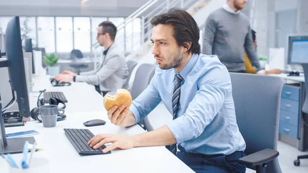 明るい現代オフィスで若いビジネスマンは、彼のデスクトップコンピュータに座ってハンバーガーを食べます,彼はパンをチューイングし、彼の昼食中に働き続けます.バックグラウンドで働く同僚 — ストック写真