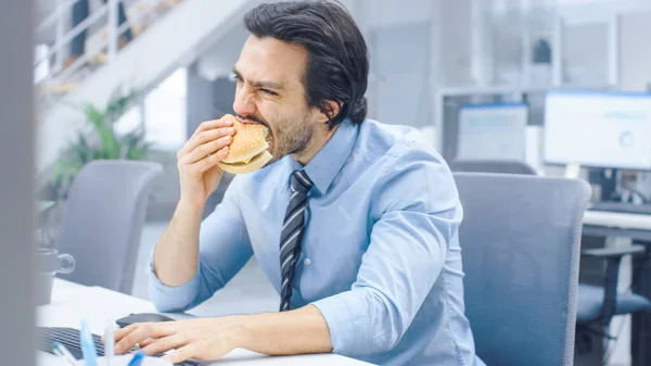 明るい現代のオフィスで若いビジネスマンは自分のデスクトップコンピュータに座ってハンバーガーを食べる,彼はパンに怒ってかむと彼の昼食中に動作し続ける.バックグラウンドで働く同僚 — ストック写真