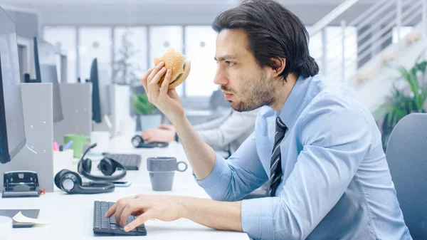 В ярком современном офисе молодой бизнесмен ест гамбургер, сидя за своим настольным компьютером, жует булочку и продолжает работать во время обеда. На заднем плане Коллеги — стоковое фото