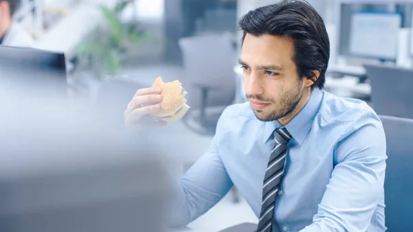 In the Bright Modern Office Young Businessman Eats Hamburger Sitting at His Desktop Computer 에서, 그는 자신의 점심 시간 동안 일을 계속하여 한다. 배경 동료들과 함께 일하면서 — 스톡 사진