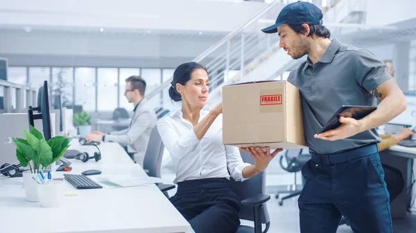 El hombre feliz de la entrega entra en oficina corporativa, paquete de las manos a una mujer de negocios hermosa. Sostiene Tablet Computer. Oficina moderna brillante grande de la compañía del negocio con los empresarios profesionales que trabajan — Foto de Stock