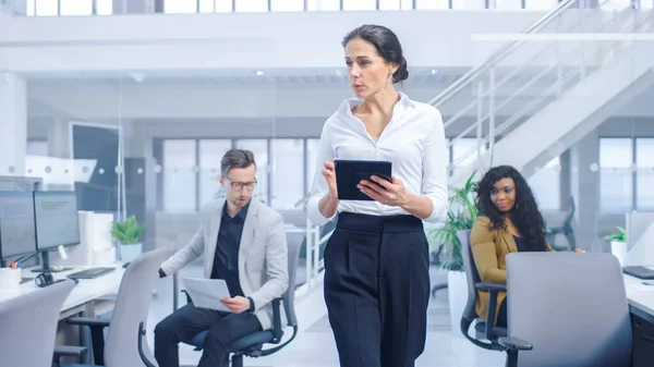 Im hellen Firmenbüro: Eine weibliche Führungskraft mit digitalen Tablet-Computern geht durch das Büro, in dem ihre professionellen Mitarbeiter an Desktop-Computern arbeiten — Stockfoto