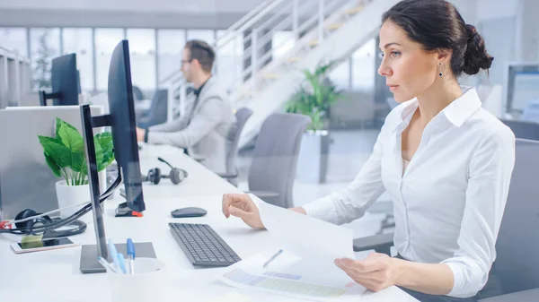 Beautiful Businesswoman, Economist, Stock Broker Uses Desktop Computer, Typing on a Keyboard, Looking At Sheets, Documents with Graphs and Statistics. in the Background Big Corporate Firm Office