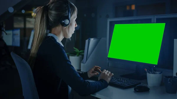 Arbeiten bis spät in die Nacht im Büro: Schöne Geschäftsfrau mit Kopfhörern und Schreibtischrechner mit grünem Bildschirm. Callcenter-Mitarbeiter, Finanzmanager, Notdienstmitarbeiter. — Stockfoto