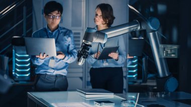 Laptop ve Tablet Analiz ile Erkek ve Kadın Mühendisler ve Bir Geleceğin Robotik Kolunun Metal Bir Cismi Nasıl Çalıştırdığını ve Taşıdığını Tartışma. Modern ekipmanlarla Yüksek Teknoloji Araştırma Laboratuvarındalar..