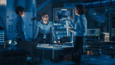 Laptop ve Tablet Analiz ve Fütürist Robotik Kol Çalışır ve Metal Cismi Taşır ile Çeşitli Mühendisler Ekibi. Modern ekipmanlarla Yüksek Teknoloji Araştırma Laboratuvarındalar..