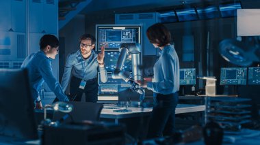 Laptop ve Tablet Analiz ve Fütürist Robotik Kol Çalışır ve Metal Cismi Taşır ile Çeşitli Mühendisler Ekibi. Modern ekipmanlarla Yüksek Teknoloji Araştırma Laboratuvarındalar..