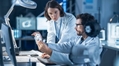 Makine Motor Geliştirme Mühendisi Masasında Bilgisayar üzerinde çalışıyor, Proje Yöneticisi ile konuşuyor, Prototip Bileşenini gösteriyor. Modern Endüstriyel Tasarım Laboratuvarı 'nda çalışan Profesyonel Ekip