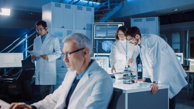 Çeşitli Uluslararası Endüstriyel Bilimadamları ve Mühendisler Takımı, Araştırma Laboratuvarında Ağır Makineler Tasarımı üzerinde çalışan beyaz önlük giyiyorlar. 3D Yazıcı, Bilgisayar ve Mikroskop kullanan Profesyoneller