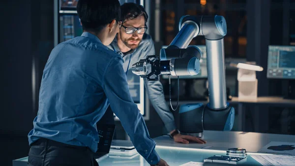 Diverso equipo de ingenieros analiza y discute cómo funciona un brazo robótico futurista y mueve un objeto metálico. Están en un laboratorio de investigación de alta tecnología con equipos modernos. — Foto de Stock