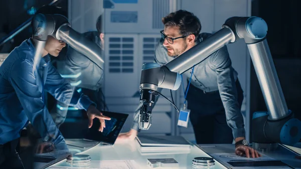 Diverso equipo de ingenieros con computadora portátil y una tableta analiza y discute cómo funciona un brazo robótico futurista y mueve un objeto metálico. Están en un laboratorio de investigación de alta tecnología con equipos modernos. —  Fotos de Stock