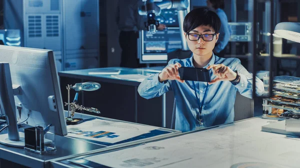 Engenheiro de Desenvolvimento Japonês Bonito em Camisa Azul está Olhando para Realirty Aumentado De Desenhos Técnicos em Seu Smartpgone no Laboratório de Pesquisa de Alta Tecnologia com Equipamento de Computador Moderno. — Fotografia de Stock