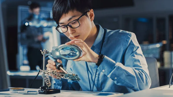 Ingénieur japonais professionnel en développement électronique en chemise bleue soude une carte de circuit dans un laboratoire de recherche de haute technologie avec un équipement informatique moderne. — Photo