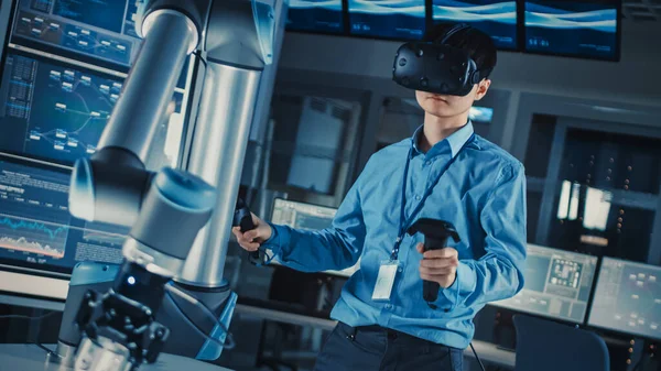 身穿蓝色衬衫的日本专业发展工程师正在用虚拟现实耳机控制一只未来派机器人臂，并在一个配备现代设备的高科技研究实验室里控制一根木棍. — 图库照片