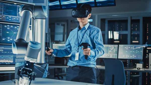 Japanischer Entwicklungsingenieur in blauem Hemd steuert einen futuristischen Roboterarm mit Virtual-Reality-Headset und Joysticks in einem High-Tech-Forschungslabor mit moderner Ausstattung. — Stockfoto