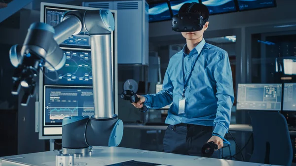 Ingegnere giapponese di sviluppo professionale in camicia blu sta controllando un braccio robotico futuristico con un auricolare di realtà virtuale e Joystick in un laboratorio di ricerca high-tech con attrezzature moderne. — Foto Stock