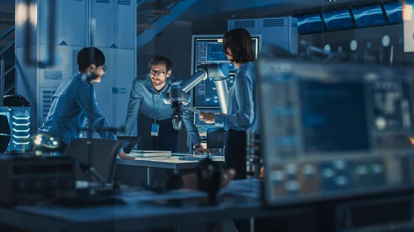 Diverso equipo de ingenieros con computadora portátil y una tableta analiza y discute cómo funciona un brazo robótico futurista y mueve un objeto metálico. Están en un laboratorio de investigación de alta tecnología con equipos modernos. —  Fotos de Stock