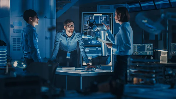 Diverso equipo de ingenieros con computadora portátil y una tableta analiza y discute cómo funciona un brazo robótico futurista y mueve un objeto metálico. Están en un laboratorio de investigación de alta tecnología con equipos modernos. —  Fotos de Stock