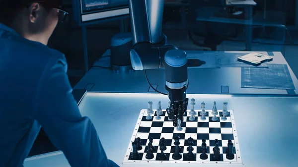 Professional Japanese Development Engineer teste une interface d'intelligence artificielle en jouant aux échecs avec un bras robotique futuriste. Ils sont dans un laboratoire de recherche moderne de haute technologie. — Photo