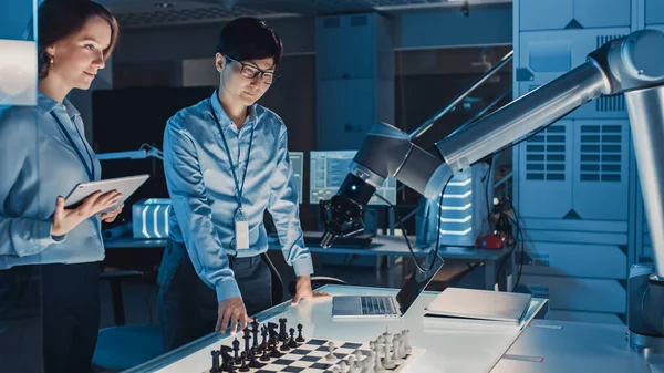 Zwei Entwicklungsingenieure diskutieren und testen eine Schnittstelle künstlicher Intelligenz, indem sie Schach mit einem futuristischen Roboterarm spielen. Sie befinden sich in einem modernen Hightech-Forschungslabor. — Stockfoto