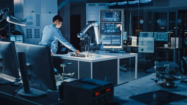 Профессиональный японский инженер-конструктор тестирует интерфейс искусственного интеллекта, играя в шахматы с футуристической роботизированной рукой. Они находятся в современной исследовательской лаборатории высоких технологий. — стоковое фото