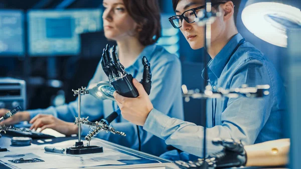 Braço de robô protético tecnológico é testado por dois engenheiros de desenvolvimento profissional em um laboratório de pesquisa de alta tecnologia com equipamentos futuristas modernos. Homem e Mulher Comparar Dados em um Pessoal — Fotografia de Stock