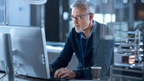 Elektroniker mittleren Alters, der in seiner Werkstatt am Personal Computer arbeitet, überprüft die Hauptplatine. Im Hintergrund verschiedene Leiterplatten, High-Tech-Geräte, Gadgets und Komponenten — Stockfoto