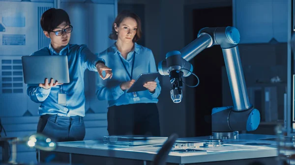 Homme et femme ingénieurs avec ordinateur portable et une tablette Analyser et discuter comment un bras robotique futuriste fonctionne et se déplace un objet métallique. Ils sont dans un laboratoire de recherche de haute technologie avec des équipements modernes. — Photo