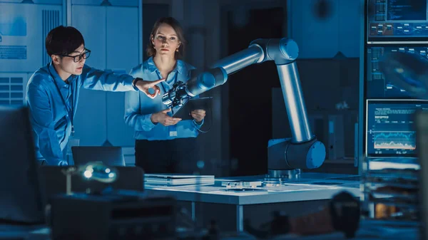 Man and Woman Engineers with Laptop and a Tablet Analyse and Discuss How a Futuristic Robotic Arm Works and Moves a Metal Object. Están en un laboratorio de investigación de alta tecnología con equipos modernos. —  Fotos de Stock