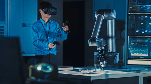Professzionális japán fejlesztő mérnök kék ingben egy futurisztikus robotkart irányít virtuális valóság headset-tel és Joysticksszel egy high-tech kutatólaboratóriumban modern berendezésekkel. — Stock Fotó