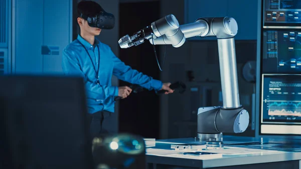 Engenheiro de desenvolvimento japonês profissional em camisa azul está controlando um braço robótico futurista com um fone de ouvido de realidade virtual e Joysticks em um laboratório de pesquisa de alta tecnologia com equipamentos modernos. — Fotografia de Stock