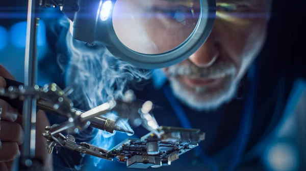 Electronics Maintenance and Repair Engineer Soldering Motherboard, Microchip and Circuit Board, Looking through Magnifying Glass, Consults Personal Computer. Close-up Low Angle Shot