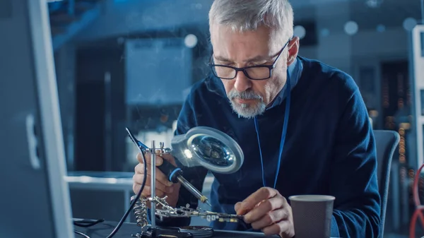 Ingeniero de mantenimiento de electrónica entra en su taller con café Beginss soldadura de placa de circuito, Mirando a través de lupa, Consulta Ordenador personal. Reparación y pruebas electrónicas — Foto de Stock