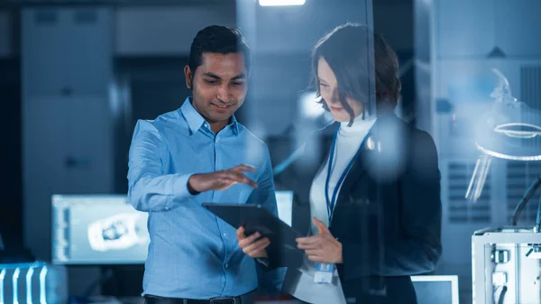 I Technology Research Facility: Kvinnlig projektledare samtalar med chefsingenjör, konsulterar de Tablet Computer. Team av industriingenjörer, utvecklare arbetar med motorkonstruktion med hjälp av datorer — Stockfoto