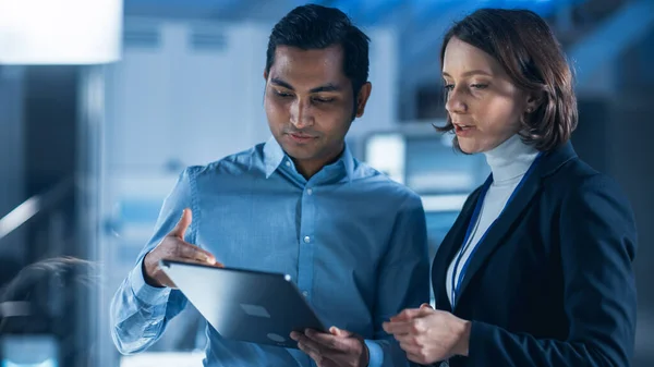 In Technology Research Facility: Female Project Manager Talks With Chief Engineer, they Consult Tablet Computer. Team of Industrial Engineers, Developers Work on Engine Design Using Computers — Stock Photo, Image