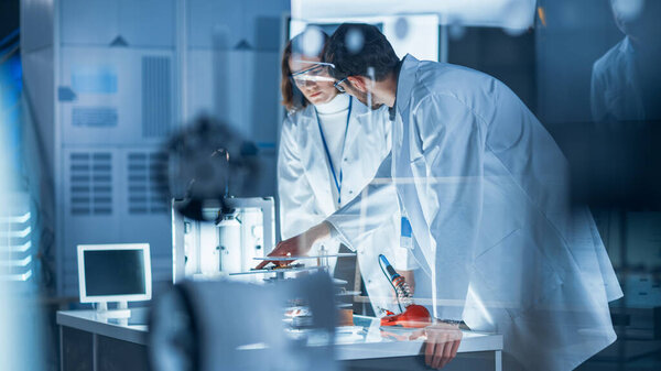 Разнообразная международная команда промышленных ученых и инженеров в белых халатах работает над проектированием тяжелого машиностроения в научно-исследовательской лаборатории. Профессионалы с помощью 3D принтера, компьютеров и микроскопа