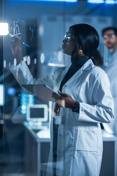 Dans le laboratoire de recherche Smart et belle scientifique afro-américaine portant un manteau blanc et lunettes de protection écrit formule sur tableau blanc en verre, Références Son ordinateur tablette — Photo