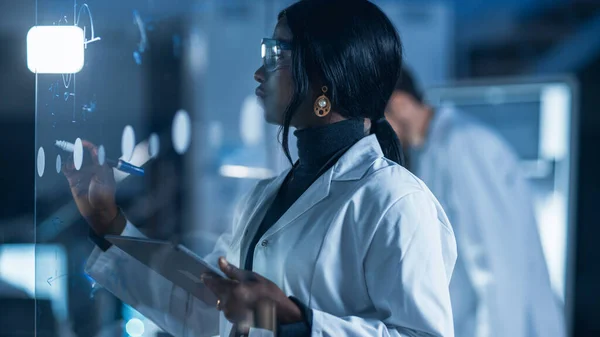 研究室でスマートと美しいアフリカ系アメリカ人女性科学者白いコートと保護ガラスを身に着けている女性科学者は、ガラスのホワイトボード上の式を書きます,参照彼女のタブレットコンピュータ — ストック写真