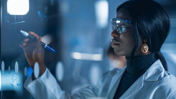 Közelkép egy okos és gyönyörű afro-amerikai tudós fehér kabátot és védőszemüveget viselő nő írja képlet üveg tábla, hivatkozás a tabletta számítógép — Stock Fotó