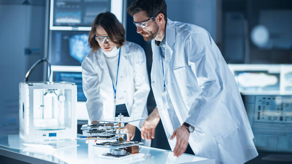 Разнообразная международная команда промышленных ученых и инженеров в белых халатах работает над проектированием тяжелого машиностроения в научно-исследовательской лаборатории. Профессионалы с помощью 3D принтера, компьютеров и микроскопа