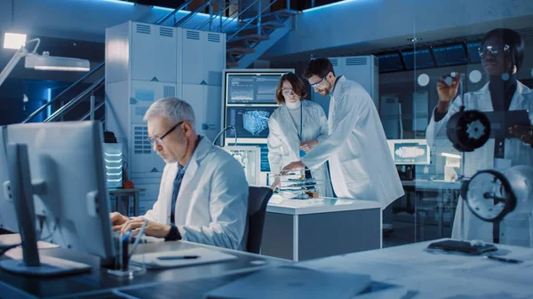 Різноманітна міжнародна команда промислових вчених та інженерів, які носять білі пальто, працюють над проектуванням важких машин у дослідницькій лабораторії. професіоналів за допомогою комп'ютерів і розмов — стокове фото
