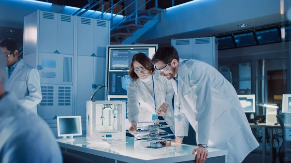 Разнообразная международная команда промышленных ученых и инженеров в белых халатах работает над проектированием тяжелого машиностроения в научно-исследовательской лаборатории. Профессионалы с помощью 3D принтера, компьютеров и микроскопа — стоковое фото
