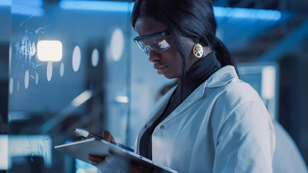 W Laboratorium Badawczym Inteligentna i Piękna Afroamerykanka Ubrana w Biały Płaszcz i Okulary Ochronne Pisze Formułę na Tablicy Szkła, Referencje Jej Komputer Tabletkowy — Zdjęcie stockowe