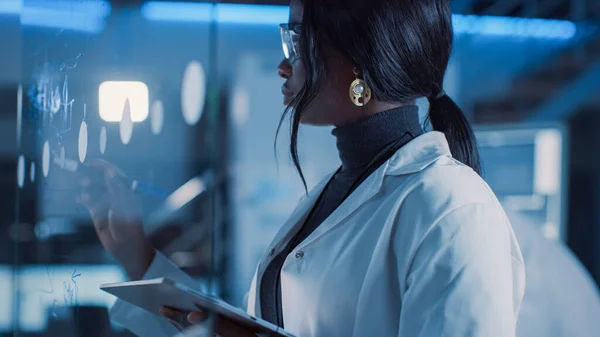 In the Research Laboratory Smart and Beautiful African American American American Scientist Wearing White Coat and Protective Glasses Writes Formula on Glass Whiteboard, References Her Tablet Computer — 스톡 사진
