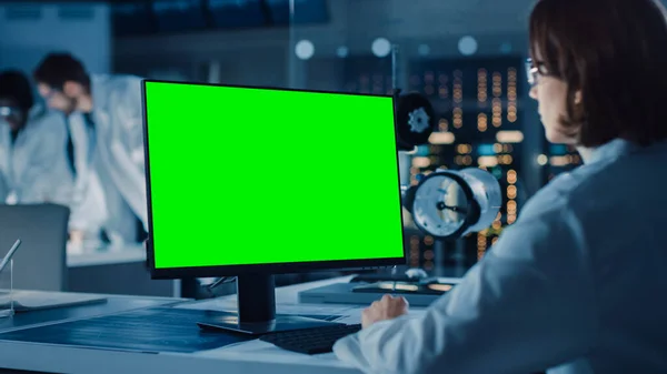 Over the Shoulder Shot: Female IT Scientist Uses Green Mock-up Screen Computer. In the Background Technology Research And Development Laboratory with Scientists, Engineers Working on High Tech Design