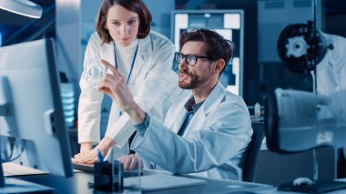 Makine Motor Geliştirme Mühendisi Masasında Bilgisayar üzerinde çalışıyor, Proje Yöneticisi ile konuşuyor, Prototip Bileşenini gösteriyor. Modern Endüstriyel Tasarım Laboratuvarı 'nda çalışan Profesyonel Ekip