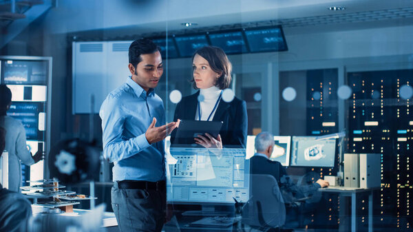 В технологическом исследовательском центре: менеджер проекта женщина беседует с главным инженером, они консультируются планшетный компьютер. Команда инженеров-промышленников, разработчиков работает над проектированием двигателей с использованием компьютеров