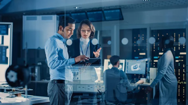 In Technology Research Facility: Female Project Manager Talks With Chief Engineer, they Consult Tablet Computer. Team of Industrial Engineers, Developers Work on Engine Design Using Computers — Stock Photo, Image