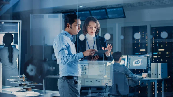 Em Technology Research Facility: Gerente de Projeto Feminino conversa com Engenheira Chefe, eles consultam o Tablet Computer. Equipe de Engenheiros Industriais, Desenvolvedores Trabalhar no Projeto do Motor Usando Computadores — Fotografia de Stock